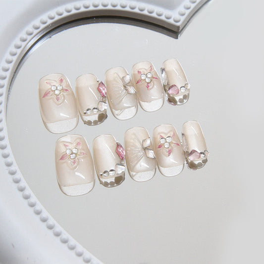 A set of beige false nails with floral patterns and rhinestone details, displayed on a white heart-shaped mirror.