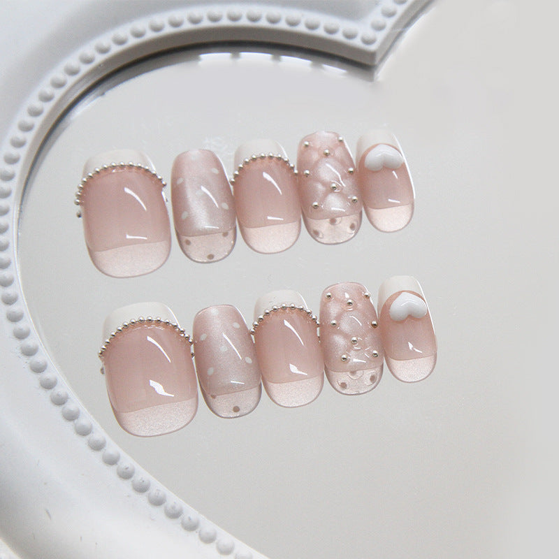 A set of long square pink press-on nails with shimmering pearls and whimsical designs, displayed on a white heart-shaped mirror.