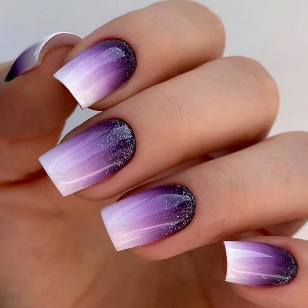 A set of press-on nails featuring a medium square gradient design with purple and white colors, accented with glitter.