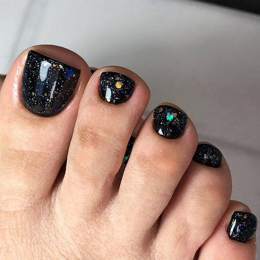 A close-up image of a person's foot with black glittery nails featuring a galaxy pattern.
