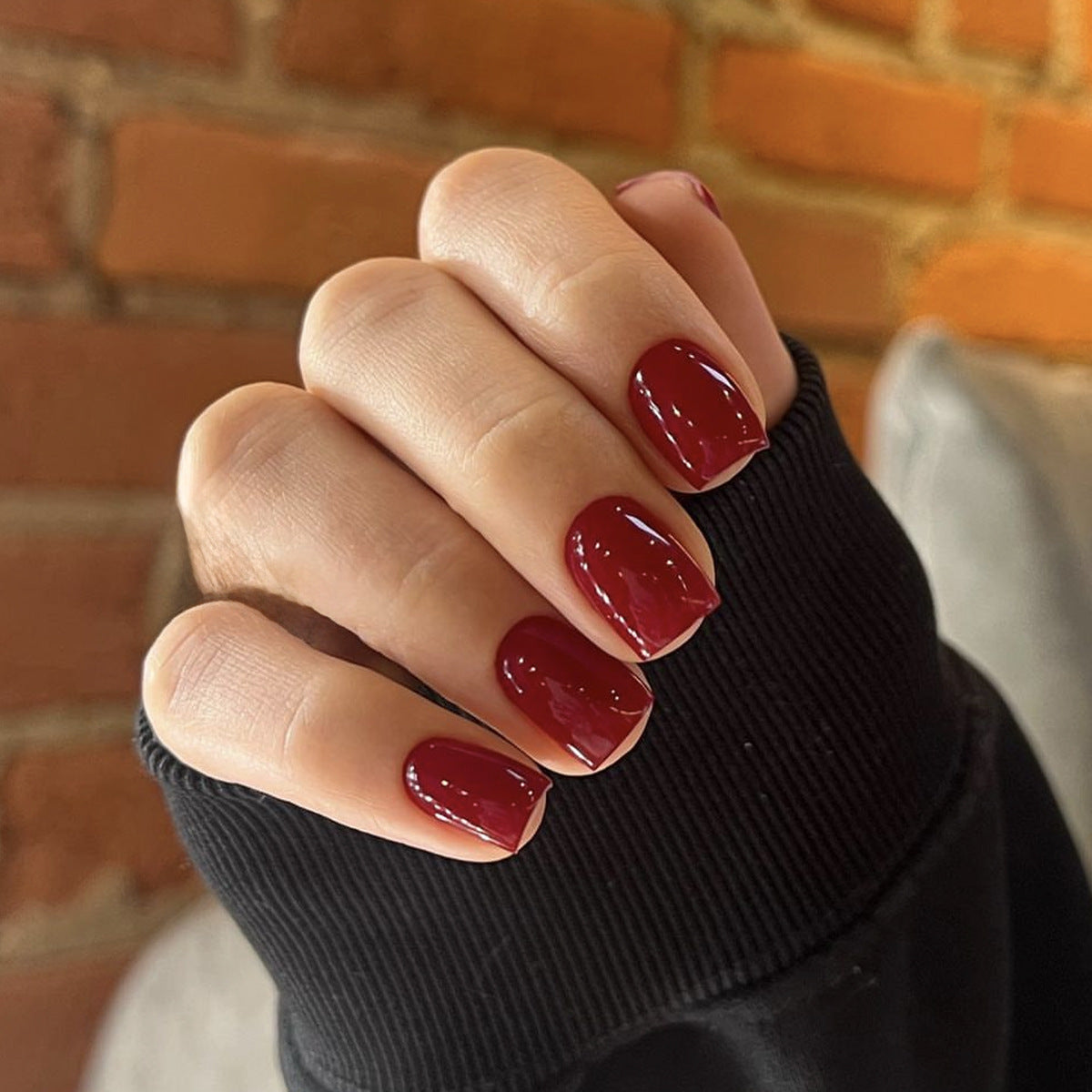 A person's hand with medium square-shaped nails, glossy burgundy red in color, resting on a textured surface with a brick wall in the background.