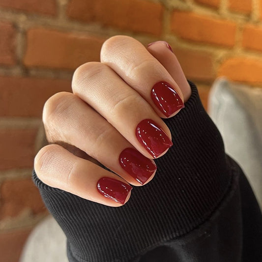 A person's hand with medium square-shaped nails, glossy burgundy red in color, resting on a textured surface with a brick wall in the background.