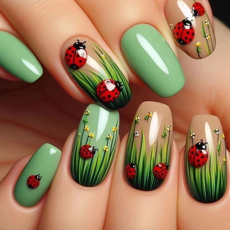 A set of press-on nails featuring a green and beige color scheme with hand-painted nature designs, including ladybugs and grass.