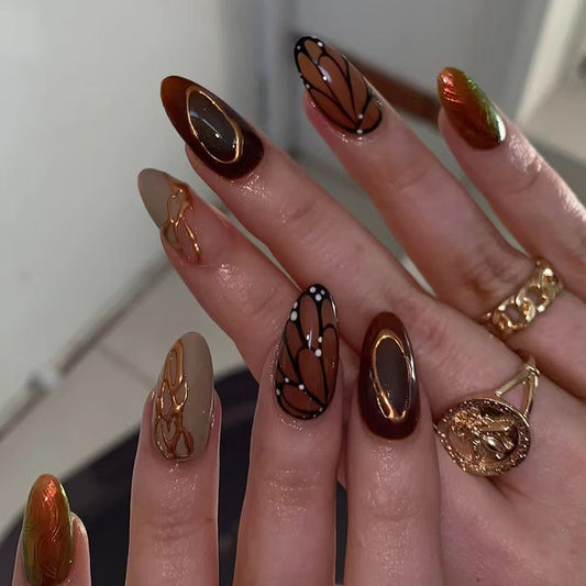 A set of long, almond-shaped brown and beige press-on nails with gold accents and unique leaf design accents.