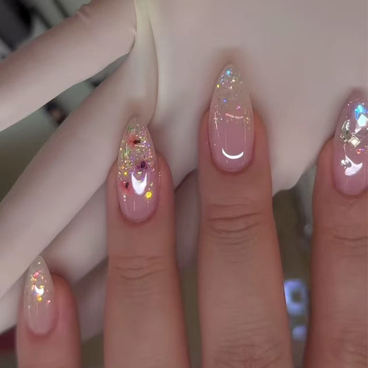 A set of long almond-shaped press-on nails with pink color and glitter, displayed on a hand wearing a glove.