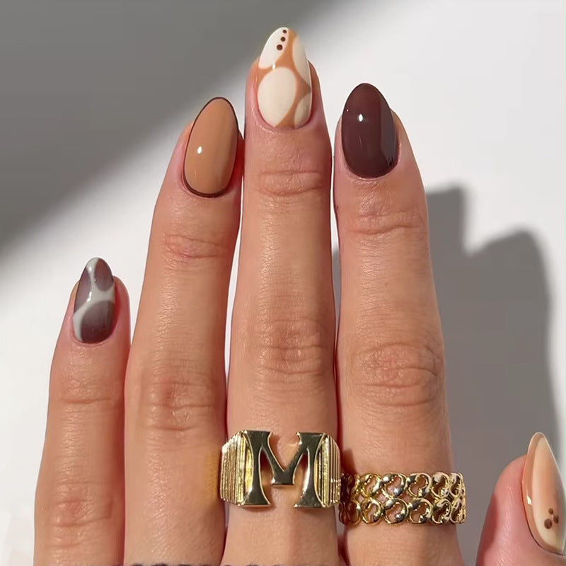 A hand with almond-shaped nails featuring a multi-tonal design with abstract patterns, displaying nail polish in shades of beige, brown, and white.