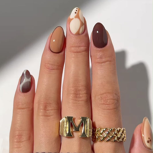 A hand with almond-shaped nails featuring a multi-tonal design with abstract patterns, displaying nail polish in shades of beige, brown, and white.