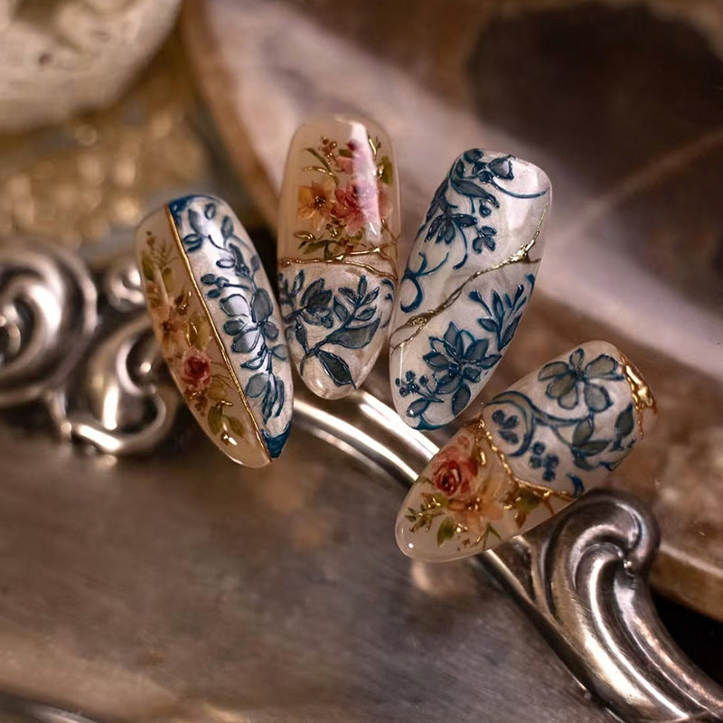 A set of almond-shaped nails with a blue and gold floral design on a metal surface.