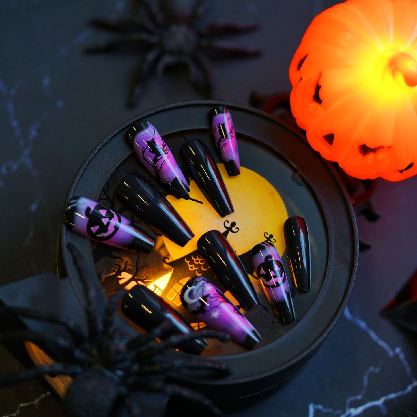 A set of long, black press-on nails with purple accents, designed with pumpkin and spider motifs, displayed in a coffin-shaped dish against a Halloween-themed backdrop with a pumpkin and spider decorations.