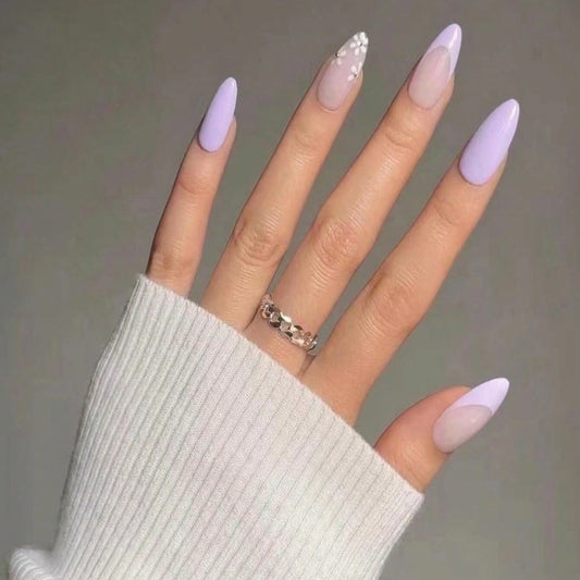 A set of long almond-shaped press-on nails in lavender color with a floral accent, shown on a hand wearing a white sweater and a ring.