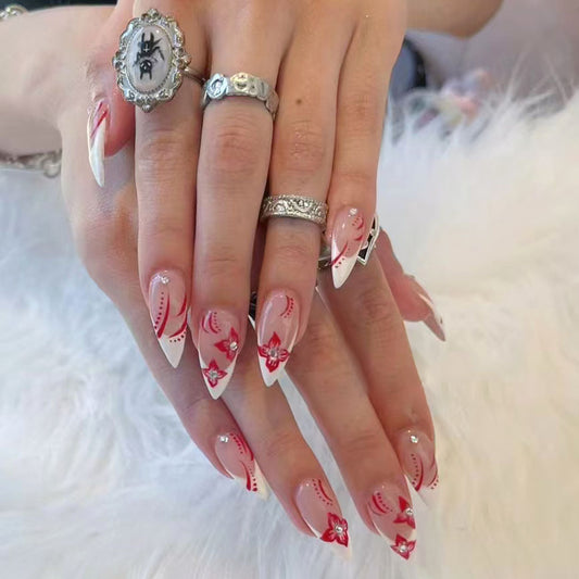 A set of long almond-shaped press-on nails with intricate red flower designs, worn on a person's hands against a white furry background.