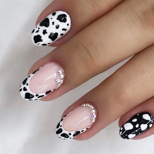 A set of press-on nails featuring black and white cattle print with rhinestone accents on a short, rounded nail shape.