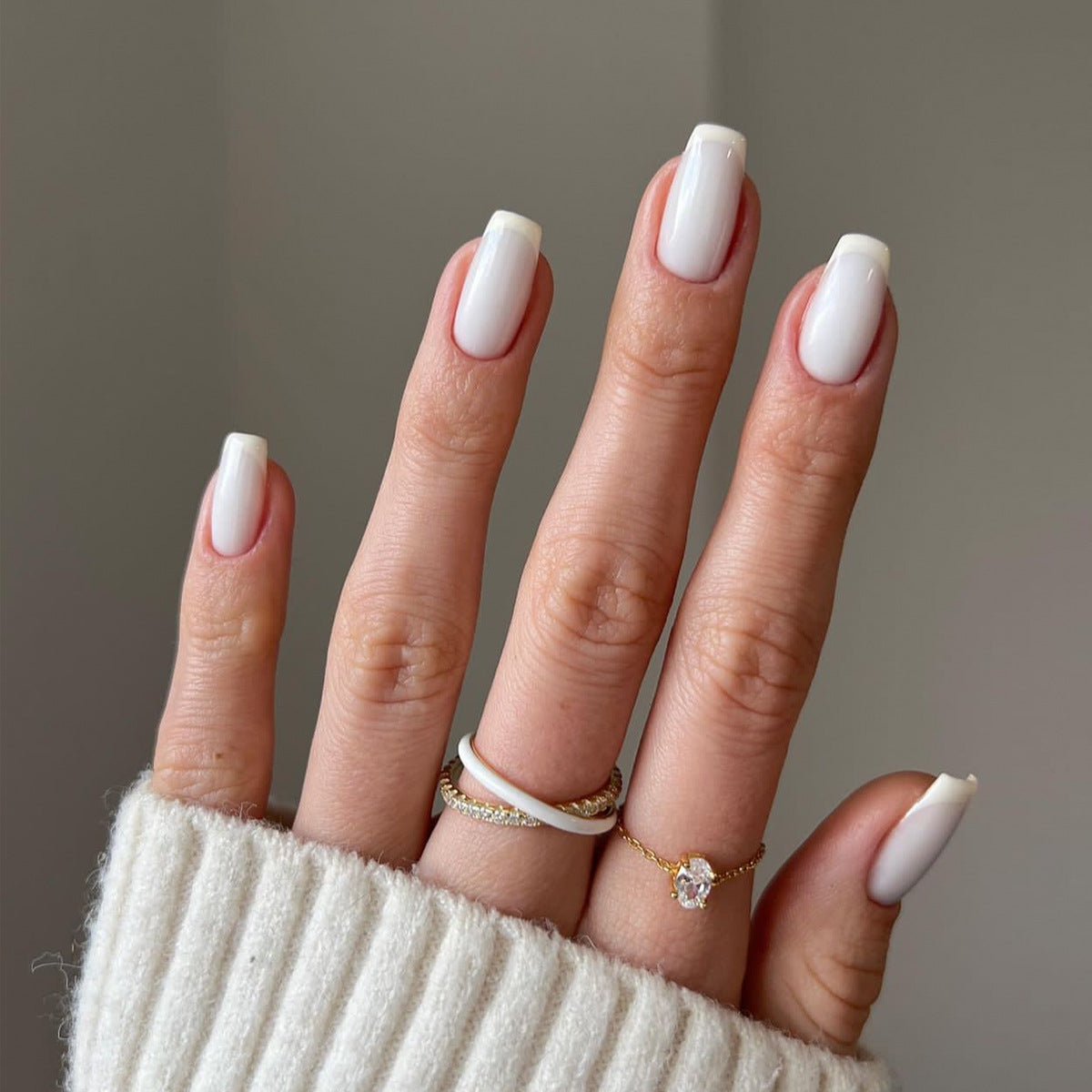 Hand with white nail polish wearing a cream sweater and gold rings on a neutral background