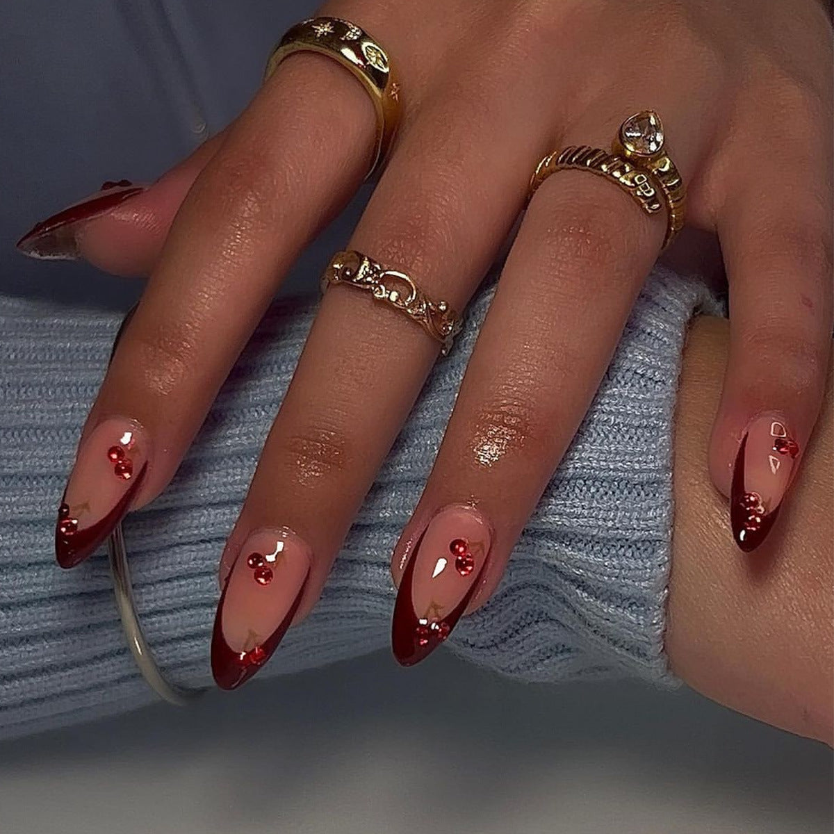 A set of long almond-shaped press-on nails with red glitter accents, displayed on a person's hand with a ring on the finger.