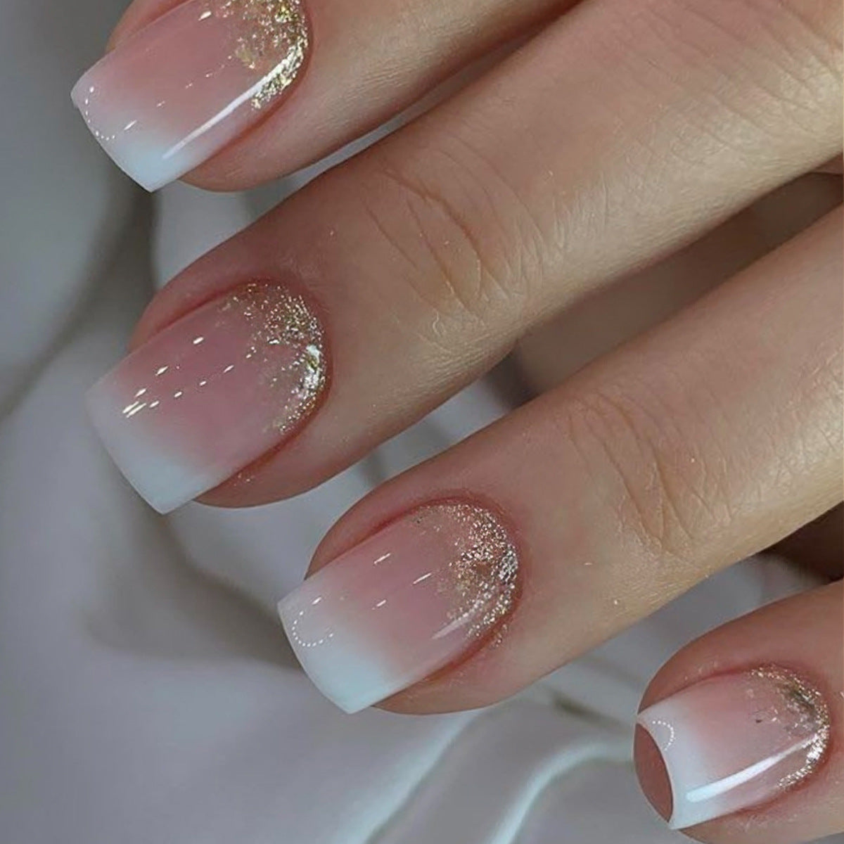 A set of press-on nails featuring a gradient of pink to white with sparkling glitter accents, presented on a hand.