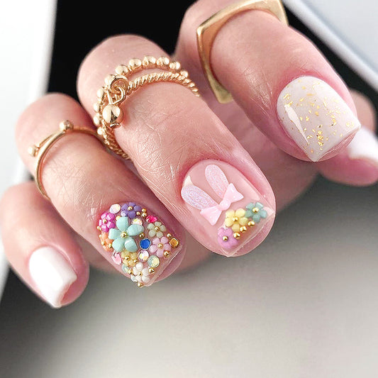 A set of press-on nails with a floral and gold flake design in pastel tones, displayed on a person's hand.
