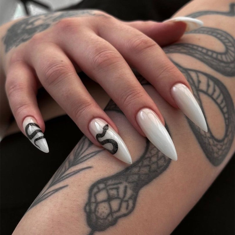 Close-up of a hand with white nail polish and black snake designs, against a tattooed arm background.