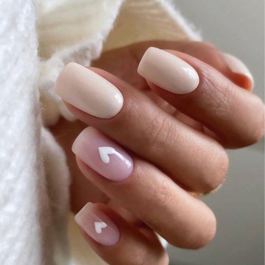 A set of pale pink press-on nails with heart accents, displayed on a person's hand.