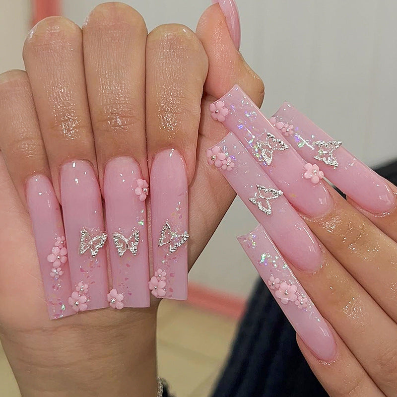 A set of long pink press-on nails with floral and butterfly accents, displayed on a person's hands.