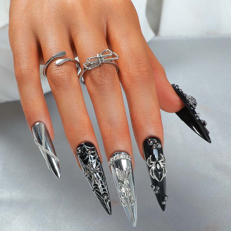 A set of long stiletto nails with intricate 3D black and silver designs, accompanied by a silver ring for attachment.