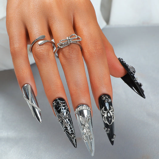 A set of long stiletto nails with intricate 3D black and silver designs, accompanied by a silver ring for attachment.