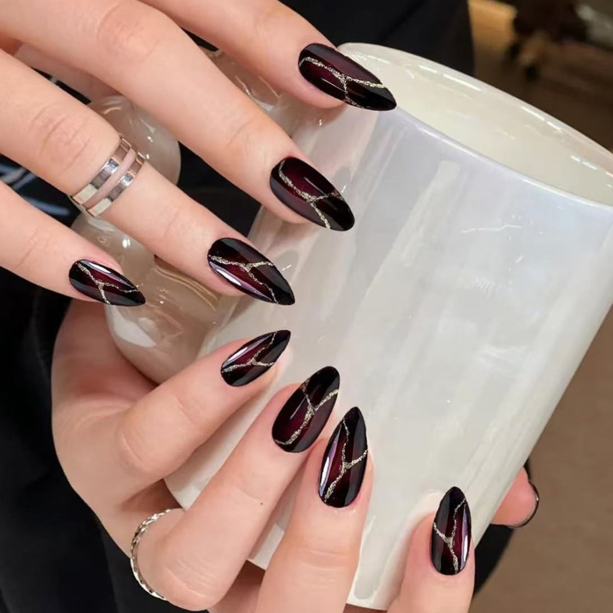 A set of almond-shaped black false nails with deep red metallic stripes, displayed on a person's fingers holding a white mug.
