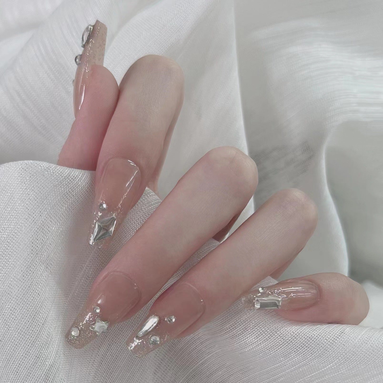 A set of long, beige-colored press-on nails with gemstone accents, displayed on a person's hand against a white textured background.
