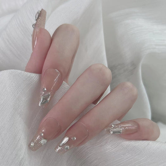 A set of long, beige-colored press-on nails with gemstone accents, displayed on a person's hand against a white textured background.