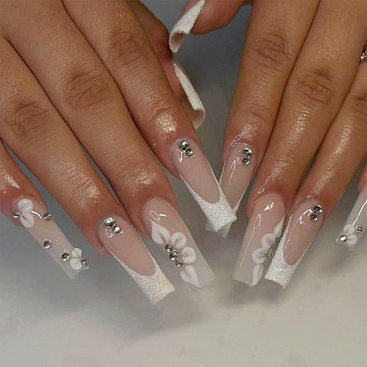 A set of long, frosted white nails with 3D flower and rhinestone accents, presented on a person's hands.