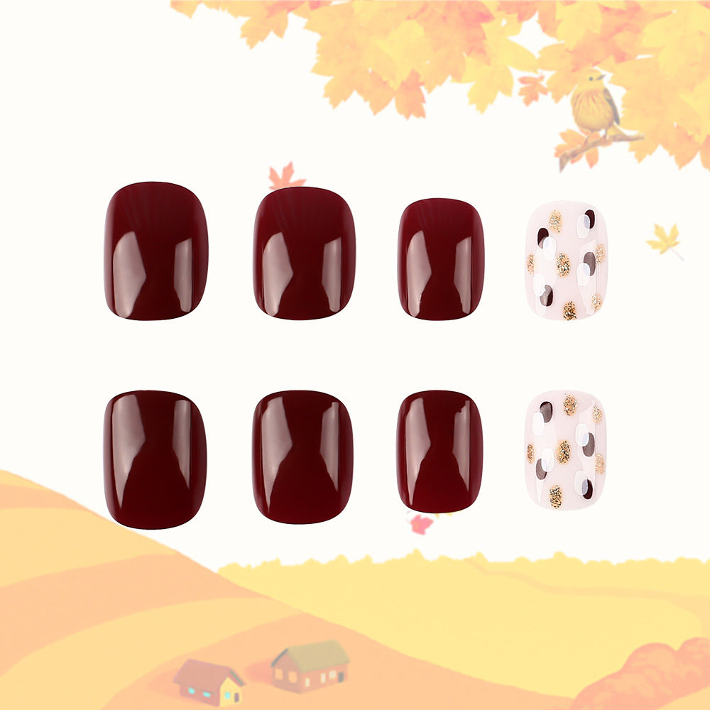 A set of four burgundy press-on nails with glitter accents, displayed with a autumn-themed background.