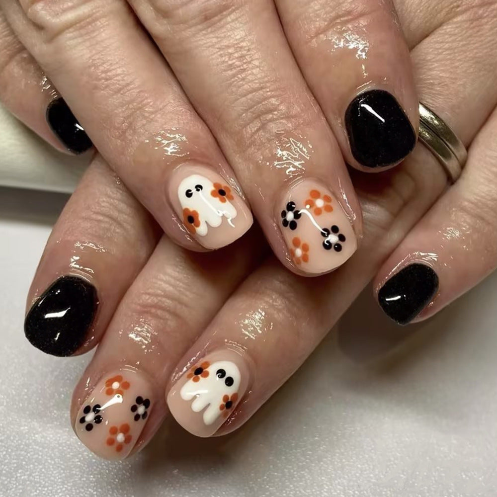A set of press-on nails featuring a black and beige color scheme with decorative elements shaped as ghosts, pumpkins, and flowers, and a glittery finish.