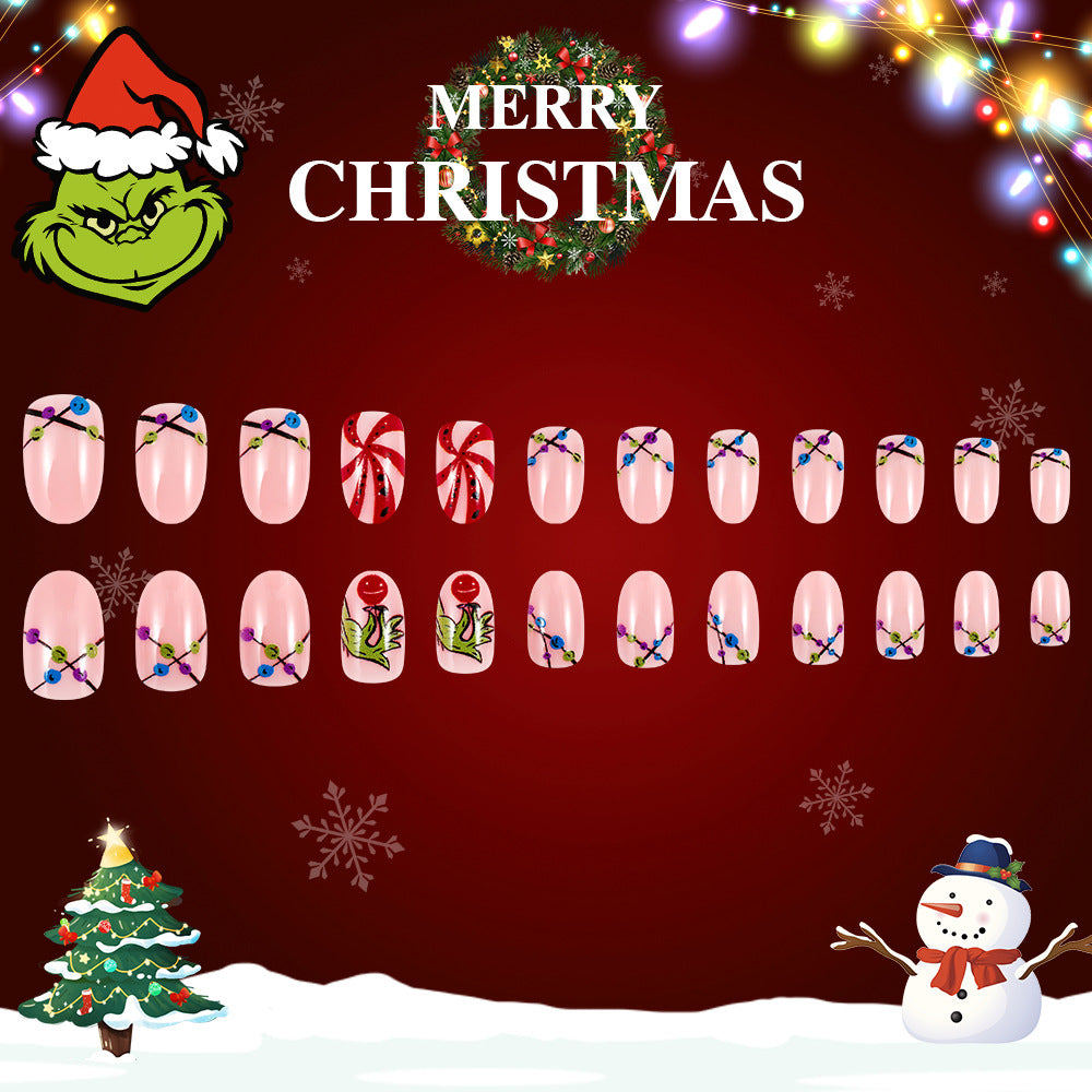 A set of press-on nails with a Christmas theme, including designs like snowflakes, ornaments, and a character resembling a grinch. The background displays a Christmas tree and snow.