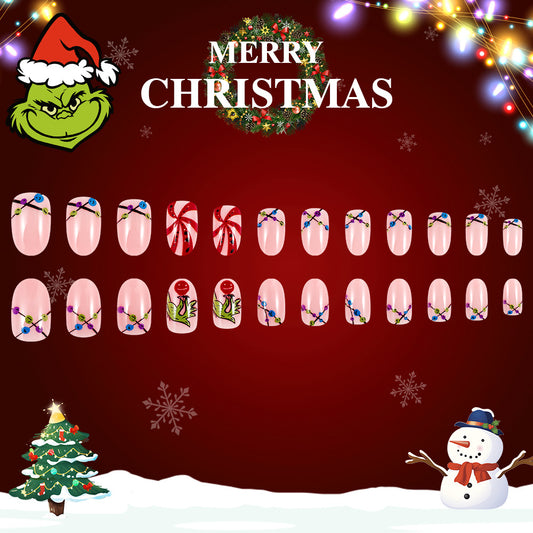 A set of press-on nails with a Christmas theme, including designs like snowflakes, ornaments, and a character resembling a grinch. The background displays a Christmas tree and snow.