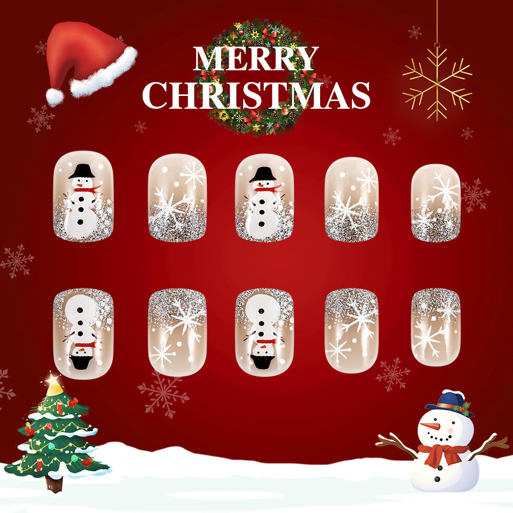 A set of press-on nails featuring a Christmas-themed design with snowman motifs in clear, white, and glitter finishes, displayed against a festive red background with snow and a Christmas tree.