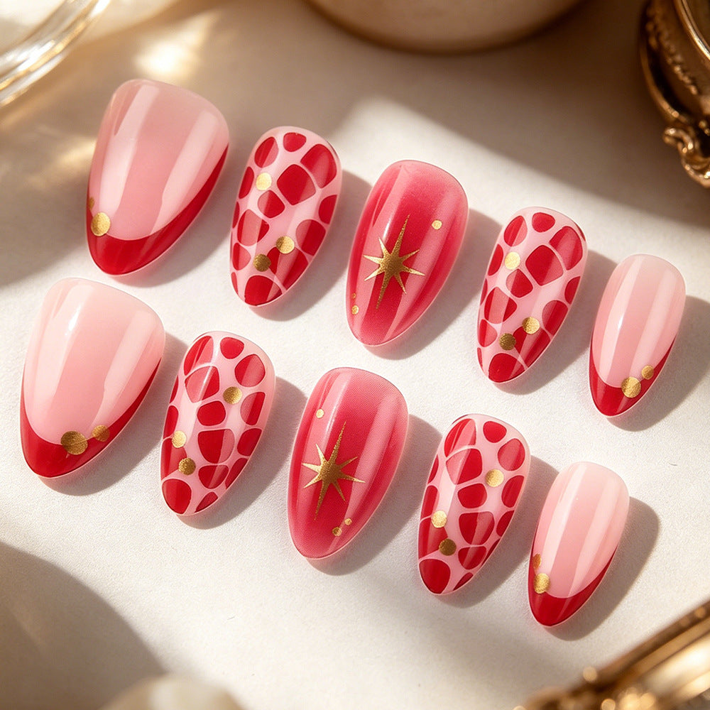 A set of artificial press-on nails arranged on a light surface. The nails feature a glossy pink and red design with some decorated in a red-and-white geometric pattern with small gold dots, and others with a red-to-pink gradient and a gold starburst motif.