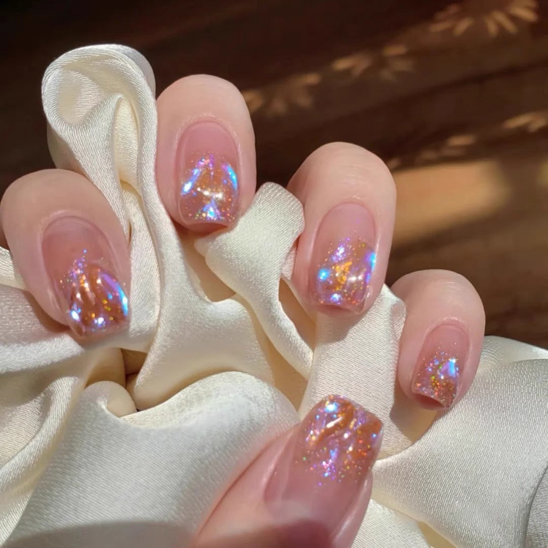 A set of short square-shaped pink nails with glitter and a holographic design, displayed on a white cloth.