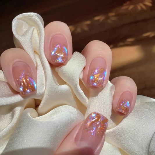 A set of short square-shaped pink nails with glitter and a holographic design, displayed on a white cloth.