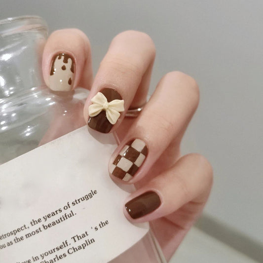 A set of brown false nails with a checkered pattern and a 3D bow accent, displayed on a person's hand.