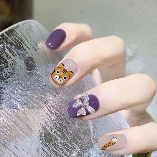 A set of purple press-on nails with glitter accents and a cartoon tiger design on one of the nails.