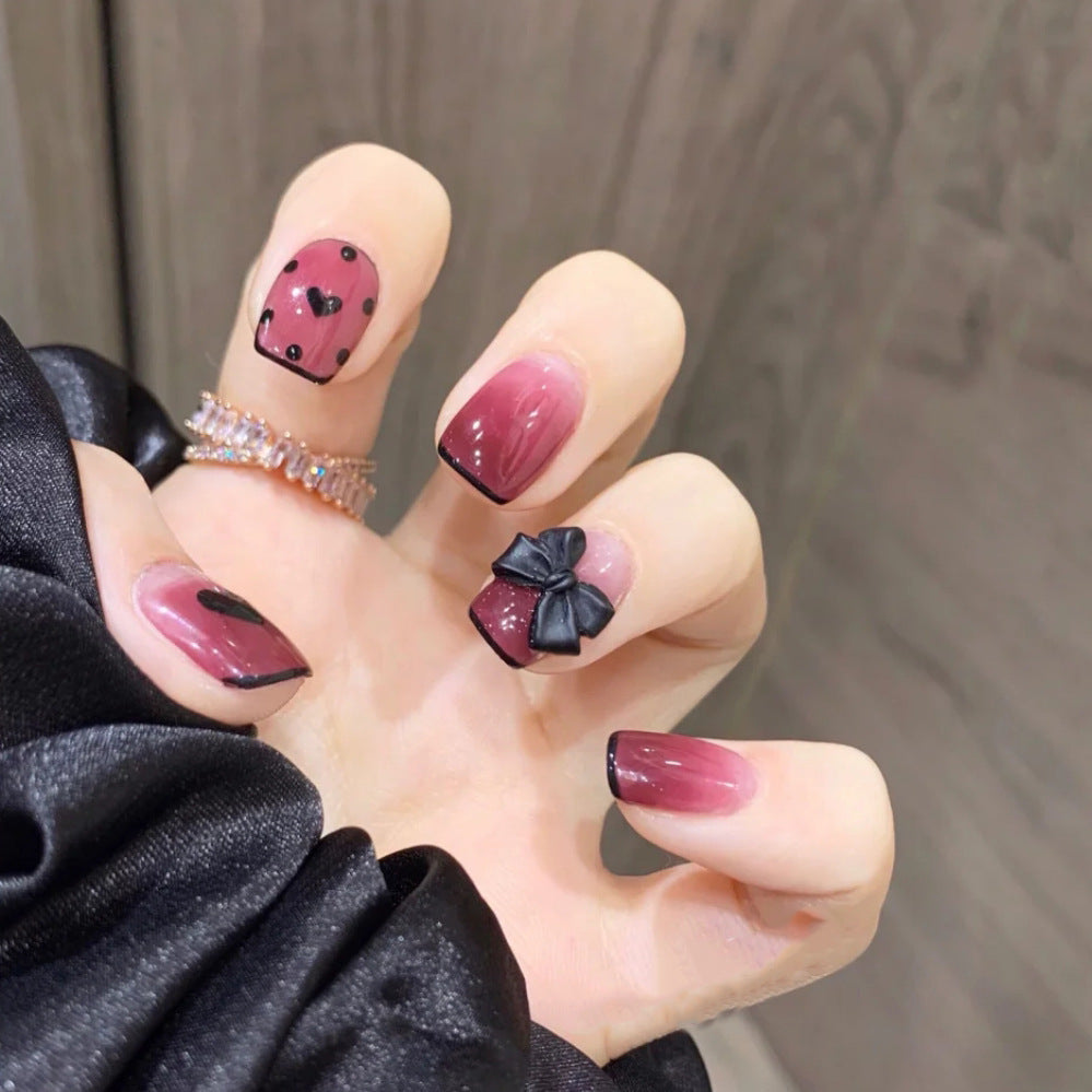 A set of burgundy and black ombre press-on nails with a 3D bow accent, displayed on a person's hand.
