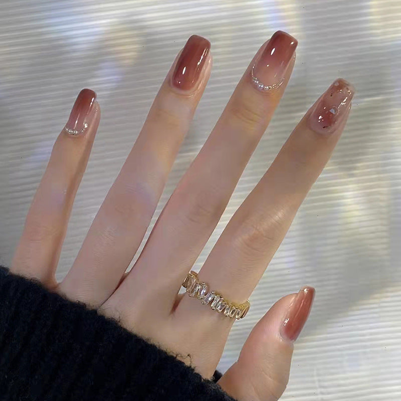 A set of taupe-colored press-on nails with rhinestone accents, displayed on a hand against a reflective background.