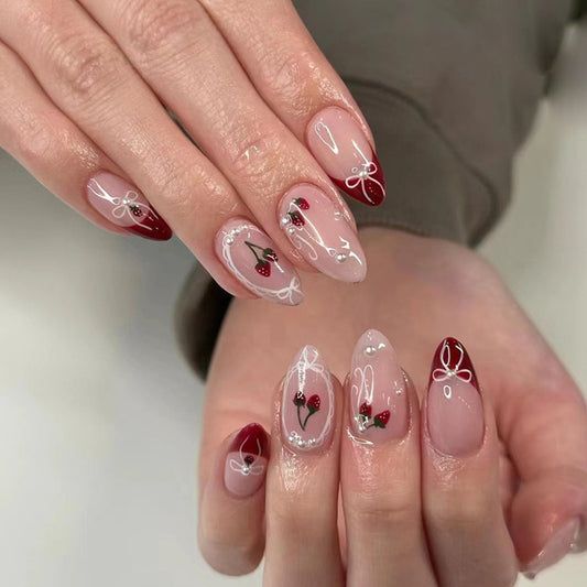 A set of press-on nails featuring medium almond shapes with a transparent base and burgundy tips, decorated with delicate floral and berry designs.