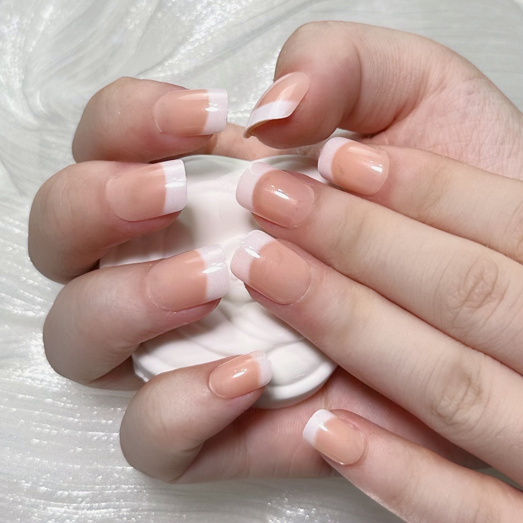 A set of long, square-shaped, beige pink and white colored press-on nails glossy finish, shown resting on a surface with a hand holding one of the nails.