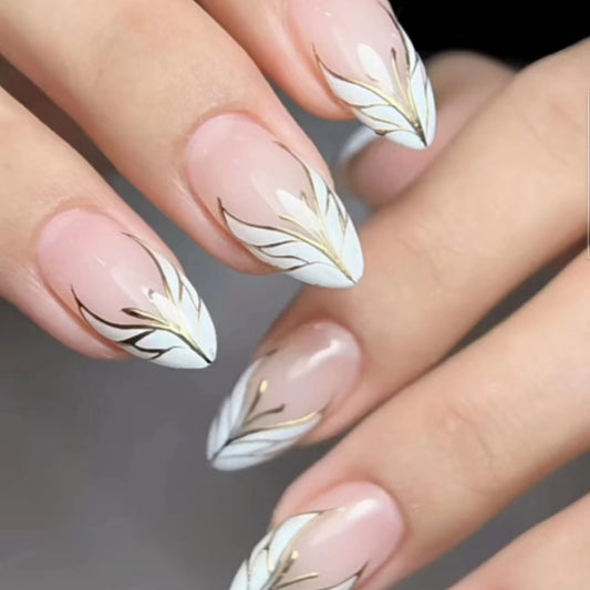 A set of almond-colored press-on nails with white and gold leaf accents, displayed on a hand.
