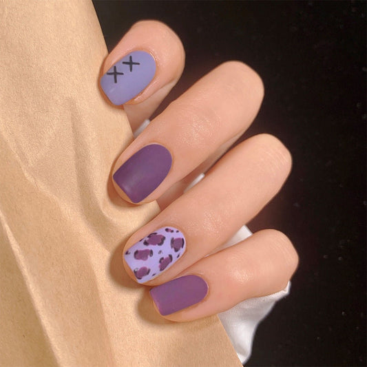 A hand with false nails painted in a matte purple color, with one nail featuring a heart pattern and another with a cross symbol.