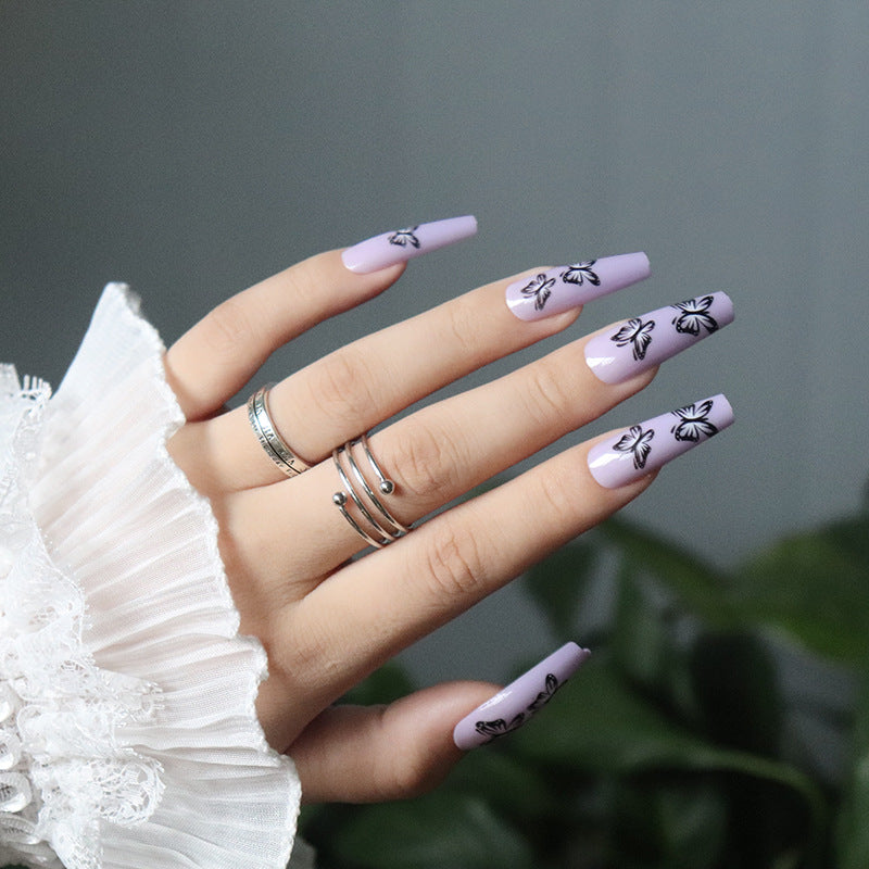 A hand with long lavender-colored press-on nails with black floral accents, displayed against a backdrop of a white ruffled sleeve and a purple-colored object in the background.