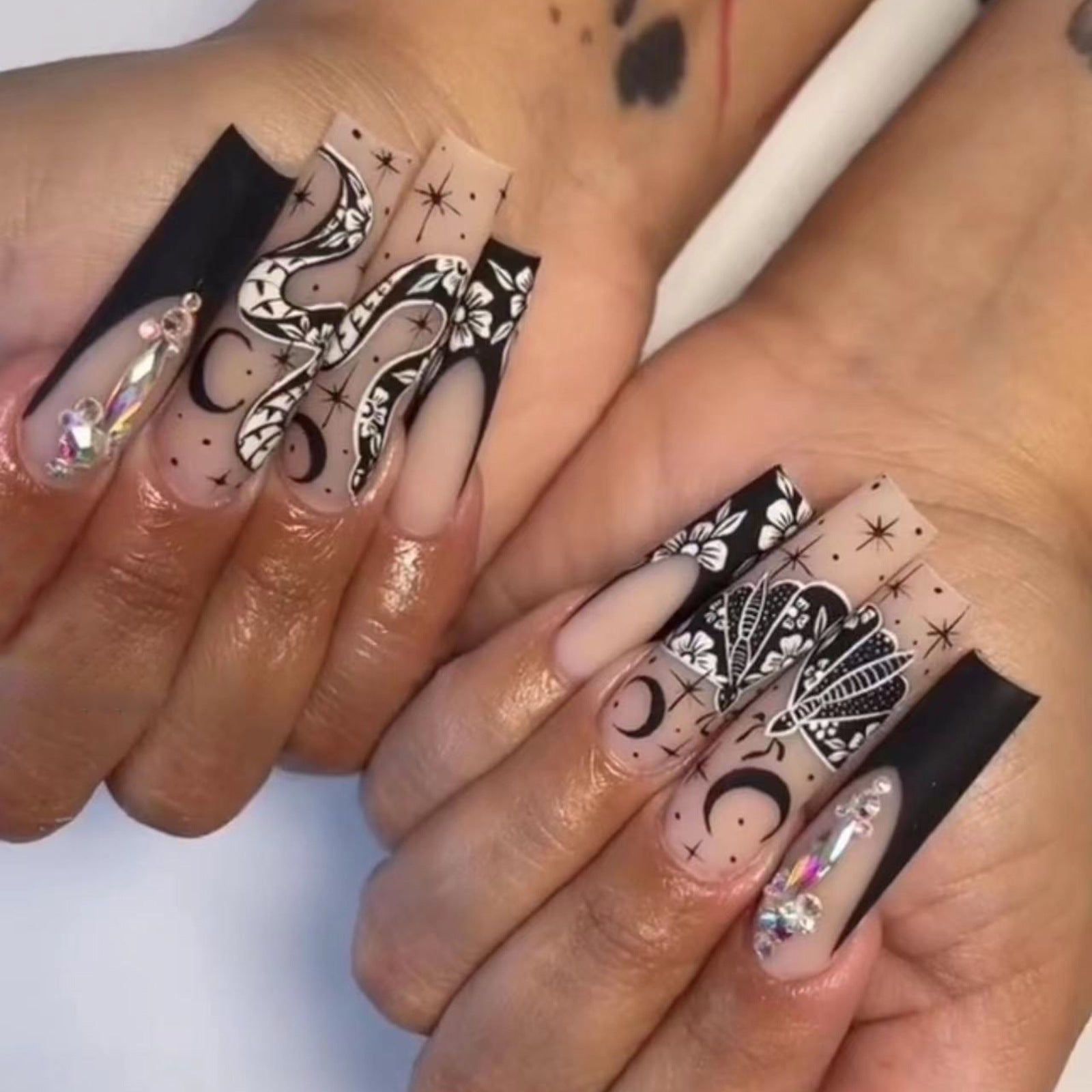 A pair of long, black, bold press-on nails featuring white and black floral and star patterns.