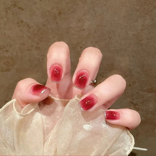 A hand with short square nails painted in red with glitter, holding a set of matching nails possibly for press-on application.