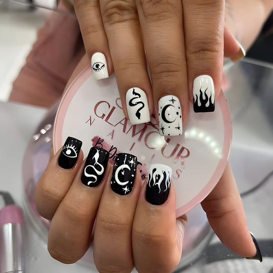 A hand showcasing press-on nails with various black and white designs, including eyes and flames, on a square black background.
