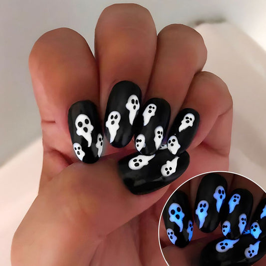 A hand showcasing press-on nails with a medium-sized, round black design featuring white ghost patterns, intended for Halloween.